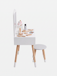 Angled side view of white wooden children's vanity with natural legs, mirror shelf, round grey stool, and scattered wooden…