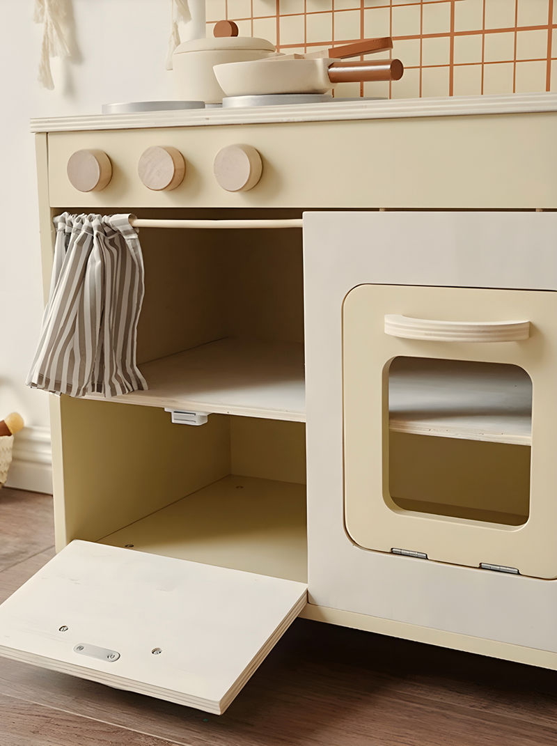 Close-up of cream wooden play kitchen lower cabinet open, striped fabric curtain, oven door ajar with window, toy pans on…
