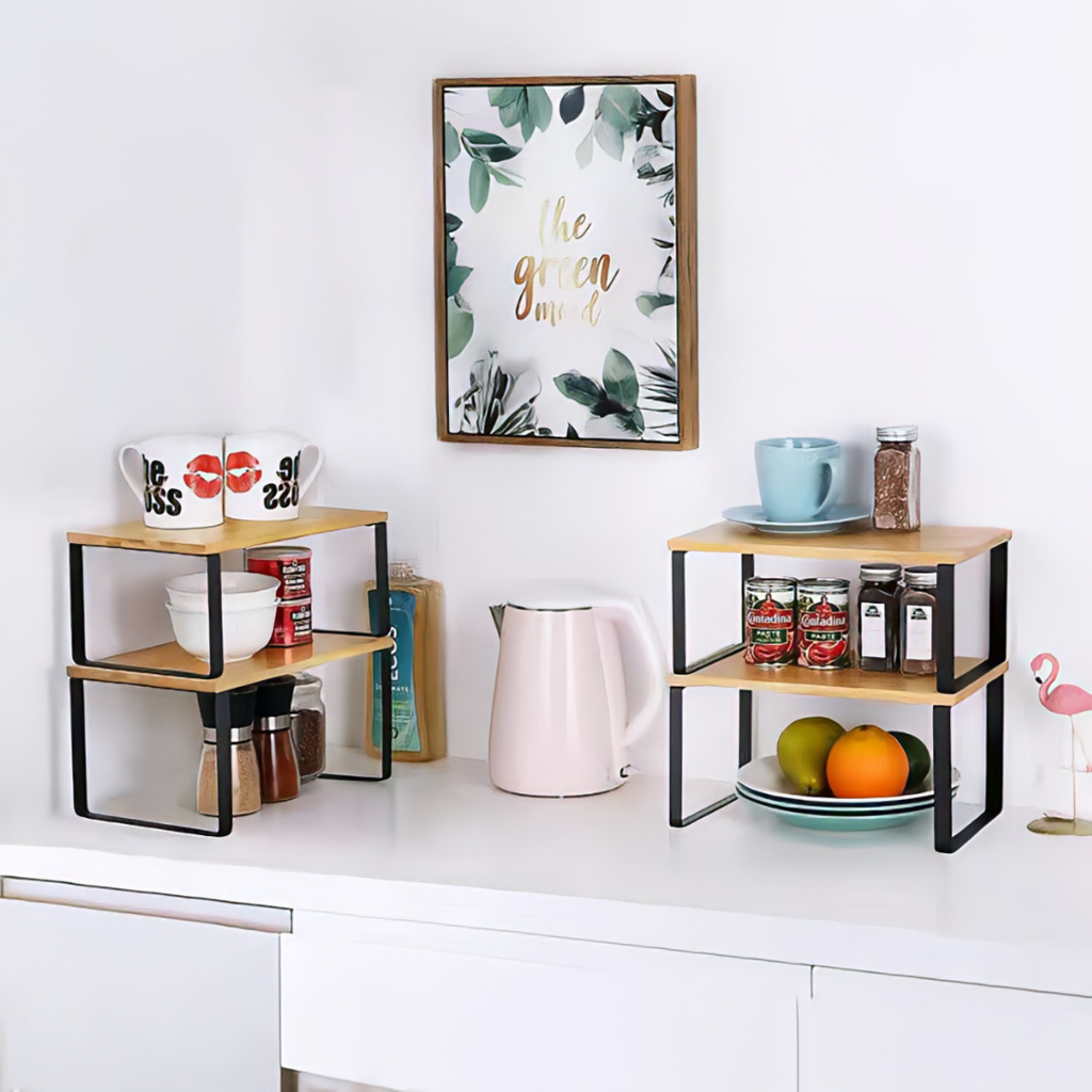 Two bamboo-top black metal counter risers on a white kitchen bench holding mugs, canned goods, spice jars, fruit, and plat…