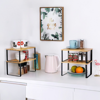 Two bamboo-top black metal counter risers on a white kitchen bench holding mugs, canned goods, spice jars, fruit, and plat…