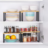 Open white kitchen cabinet with two bamboo-top black metal risers organising bowls, coloured plates, spice jars, and canne…