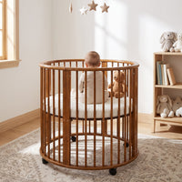 Walnut-finish round wooden baby playpen on castors, rear view with seated baby and teddy bear inside, star mobile above, n…