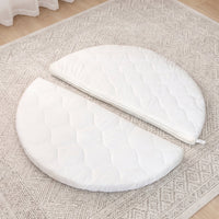 White quilted round baby mattress split into two halves, laid flat on a neutral patterned rug, overhead angle