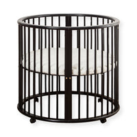 Black round wooden baby playpen with vertical spindles, white round mattress inside, and small wheels on base, white backg…