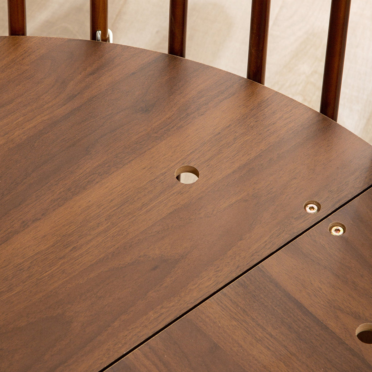 Close-up of walnut-finish round baby playpen base showing pre-drilled holes, exposed bolt fixings, and a panel join seam