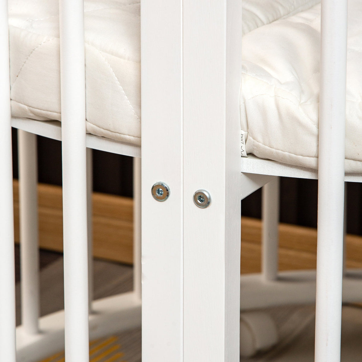 Close-up of white wooden cot frame joints secured with two bolts, cream quilted mattress visible between vertical slats