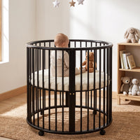 Black round wooden playpen with baby and teddy bear inside, styled in a sunlit nursery with jute rug and toy shelf nearby