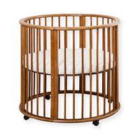 Round walnut-finish wooden baby playpen with vertical slats, white circular mattress inside, and black caster wheels, fron…