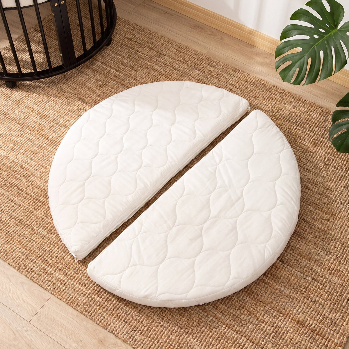 White quilted round mattress split into two halves, laid flat on a jute rug beside a black round wooden playpen
