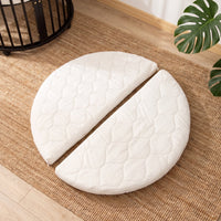 White quilted round mattress split into two halves, laid flat on a jute rug beside a black round wooden playpen
