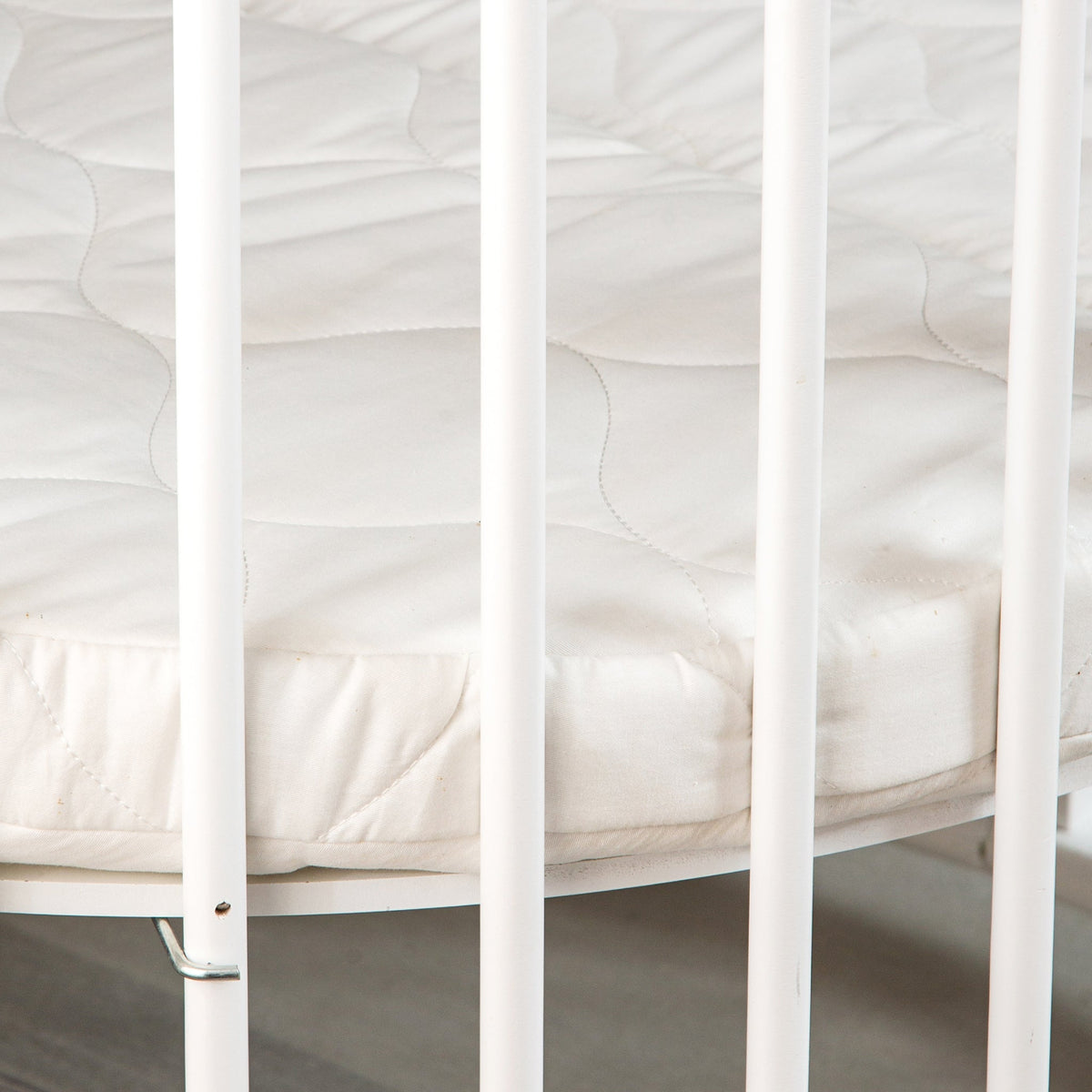 Close-up of quilted white round mattress inside white-barred baby playpen, showing mattress edge and fabric underside detail