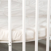 Close-up of quilted white round mattress inside white-barred baby playpen, showing mattress edge and fabric underside detail