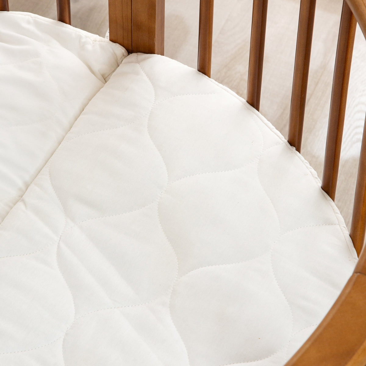 Overhead angle of a white quilted oval crib mattress fitted inside a walnut-finish round wooden baby playpen, curved slats…