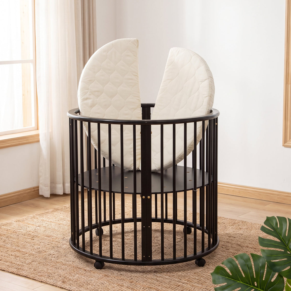 Black round wooden baby playpen on jute rug, split oval mattress propped upright inside, near sunlit window with sheer cur…
