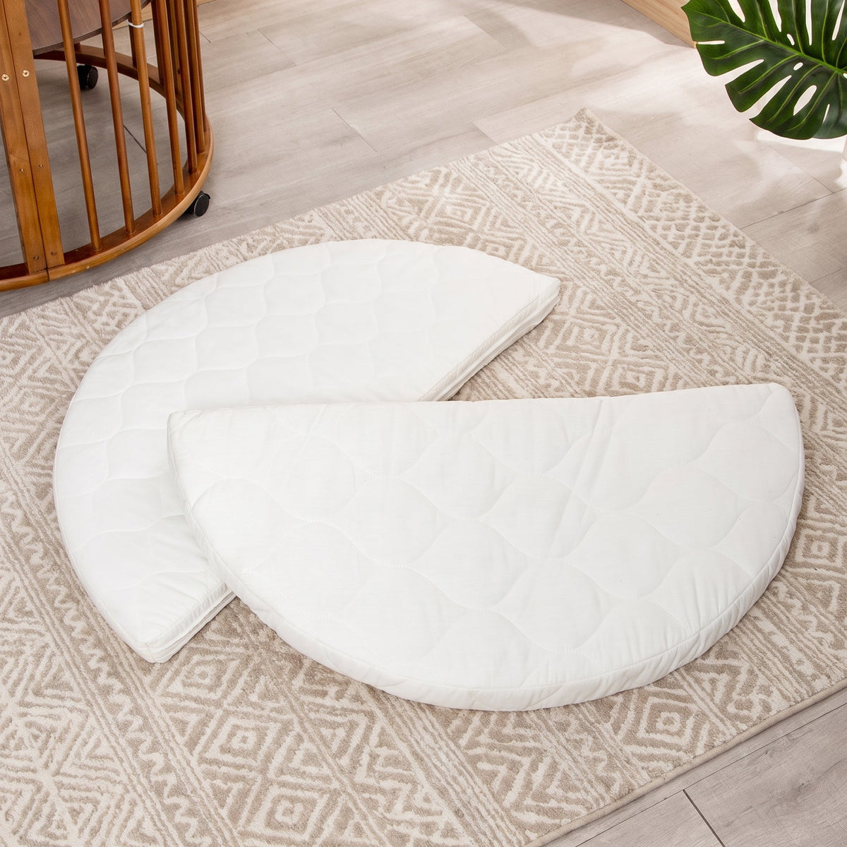 Two white quilted semicircular mattress pieces laid flat on a beige patterned rug, round wooden crib visible beside them