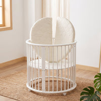 White oval wooden baby playpen on castors with cream quilted split mattress standing upright inside, on jute rug by window
