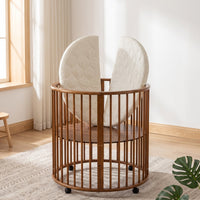 Walnut-finish round wooden baby playpen on castors, quilted oval mattress propped open vertically inside, nursery lifestyl…