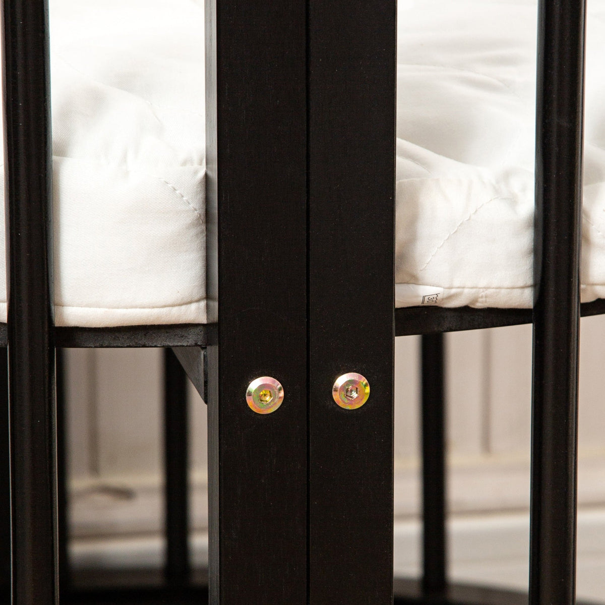 Close-up of two gold bolts securing the black wooden central post of a round baby playpen, white quilted mattress visible…