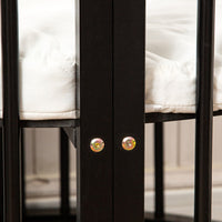 Close-up of two gold bolts securing the black wooden central post of a round baby playpen, white quilted mattress visible…