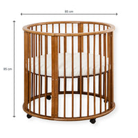 Walnut-finish round wooden baby playpen with dimension lines showing 85 cm width and 85 cm height, white mattress inside,…